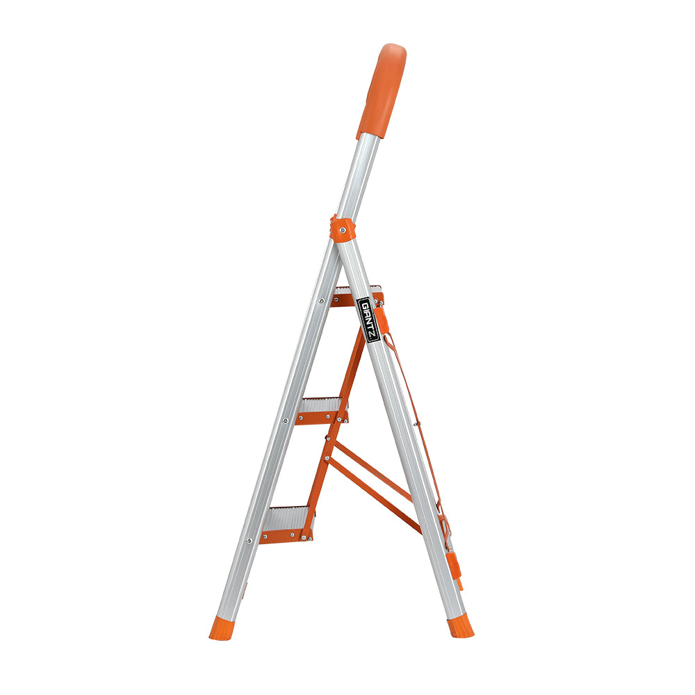 A Giantz 3 Step Ladder Multi-Purpose Folding Aluminium Light Weight Non Slip Platform, accented with orange on the steps, top railing, and foot pads. Designed for indoor use, it features non-slip rubber feet for stability and a handle at the top for added safety.