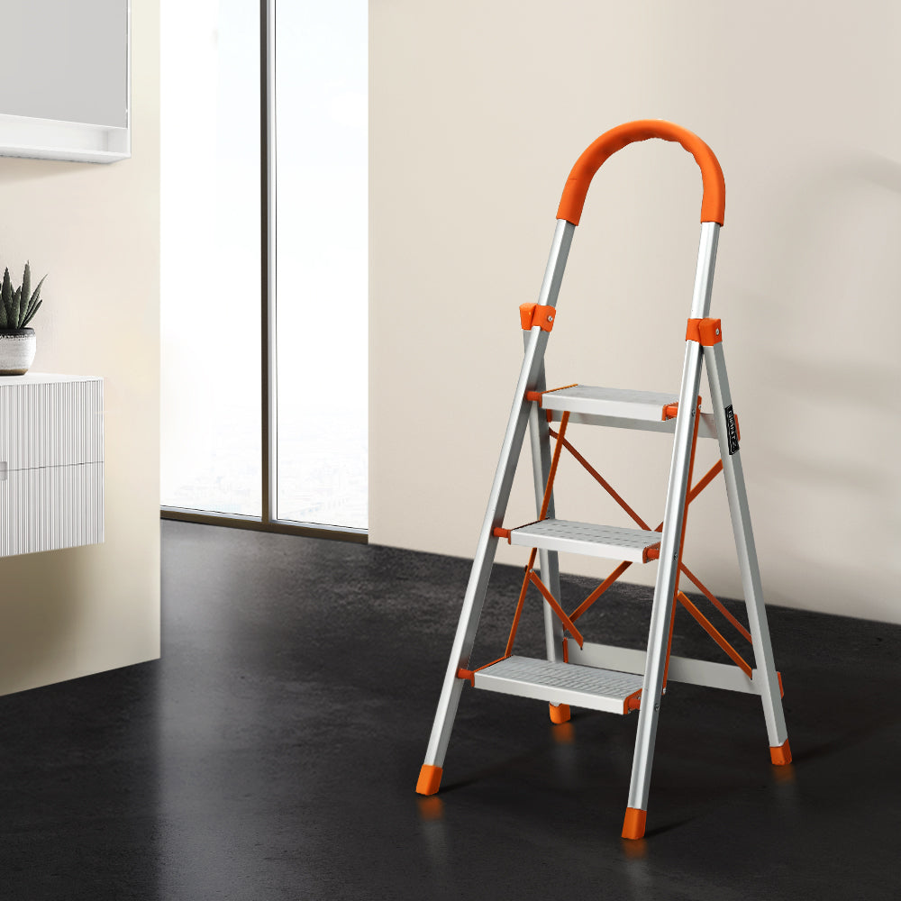 A Giantz 3 Step Ladder Multi-Purpose Folding Aluminium Light Weight Non Slip Platform, accented with orange on the steps, top railing, and foot pads. Designed for indoor use, it features non-slip rubber feet for stability and a handle at the top for added safety.