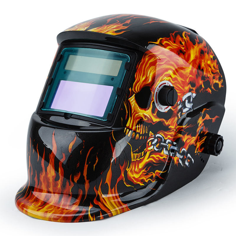 A black ROSSI Solar Auto Darkening Welding Helmet Mask MIG/ARC/TIG Welder Machine with an artistic design featuring vibrant flames and a fiery skull on the sides. This solar helmet boasts a wide viewing window with automatic tinting and an adjustable knob for optimal comfort.